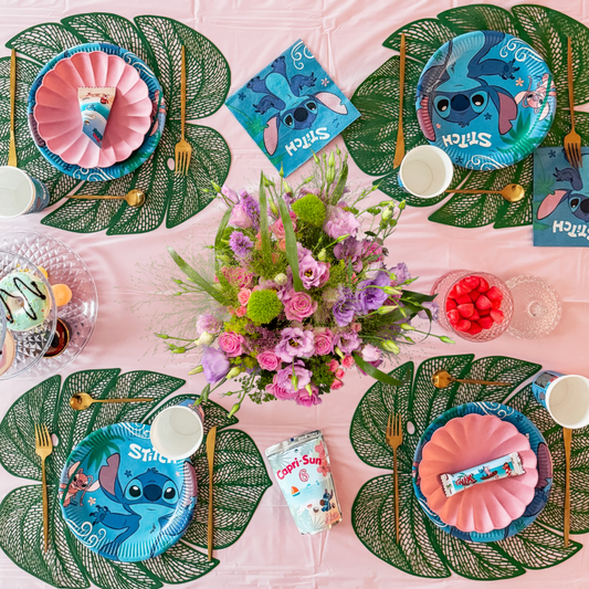 Décoration d'anniversaire Stitch pour enfant avec assiettes, serviettes, accessoires et fleurs sur une table tropicale rose