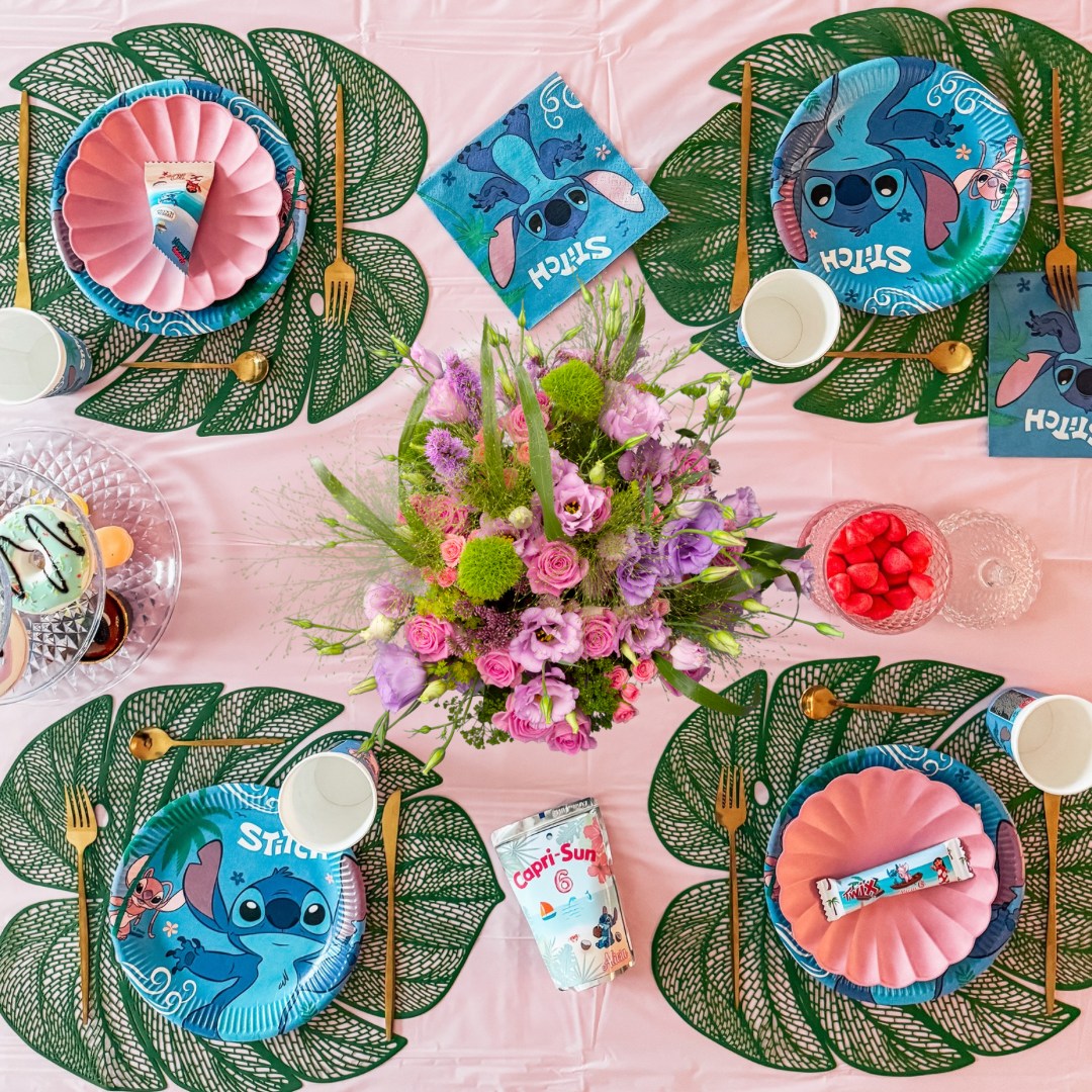 Décoration d'anniversaire Stitch pour enfant avec assiettes, serviettes, accessoires et fleurs sur une table tropicale rose